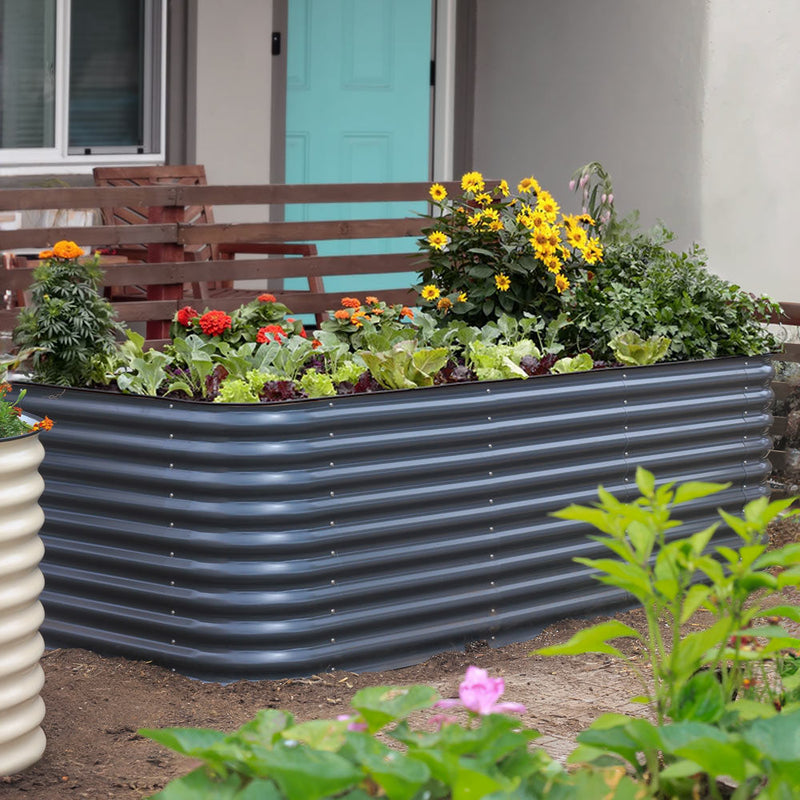 The 29-inch tall large epic metal raised garden bed filled with green plants, yellow flowers, and red flowers, situated in a garden in front of a house with a turquoise door.