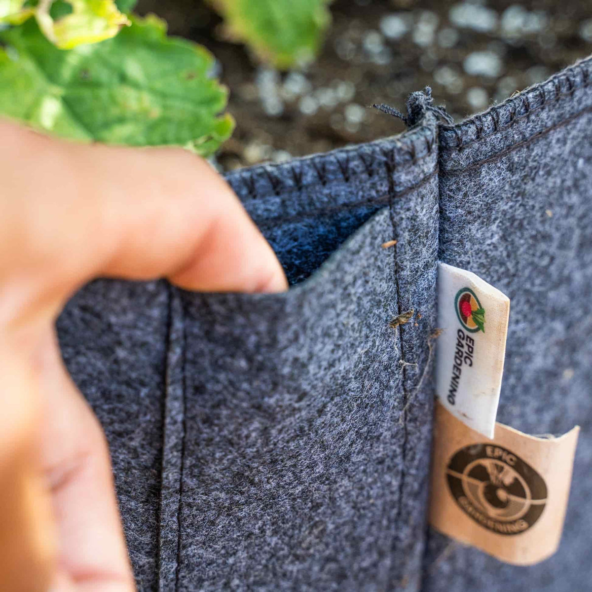 Close-up of a hand opening the reinforced stitched seam of a grey felt Epic Gardening grow bag, revealing its lined interior, with the brand's fabric and cardboard tags visible and leafy plants blurred in the background.