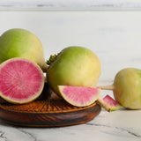 Whole watermelon radishes with pale green-and-white skin arranged on a wooden dish, with one halved and one wedge cut open to reveal vivid pink, radiating flesh inside.