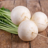 Three round, smooth white turnips with tapering root tips and their leafy green tops attached, resting on a rustic wooden surface.