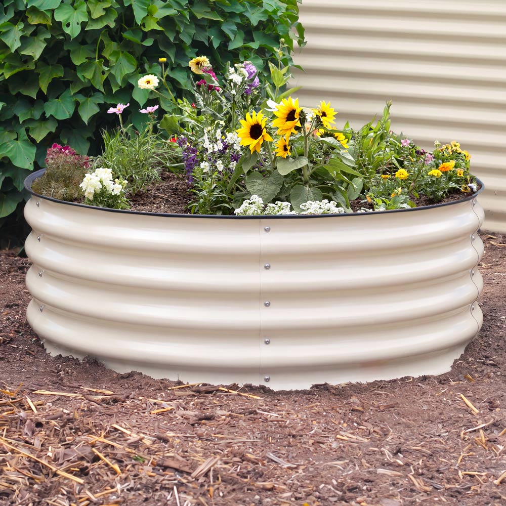A round, ribbed light clay metal raised garden bed filled with sunflowers and other colorful garden plants, set in a mulch bed against a beige wall and green foliage.