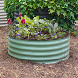 A round corrugated mist green metal raised garden bed filled with green plants, small purple flowers, and red blooms, set in a garden bed of soil mulch with leafy greenery in the background.