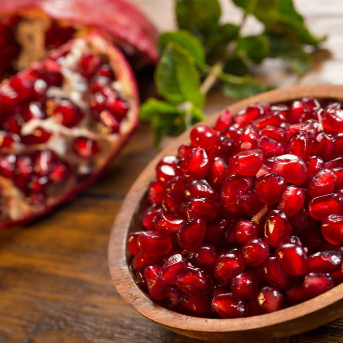 Wooden bowl filled with fresh red pomegranate arils next to a halved pomegranate.