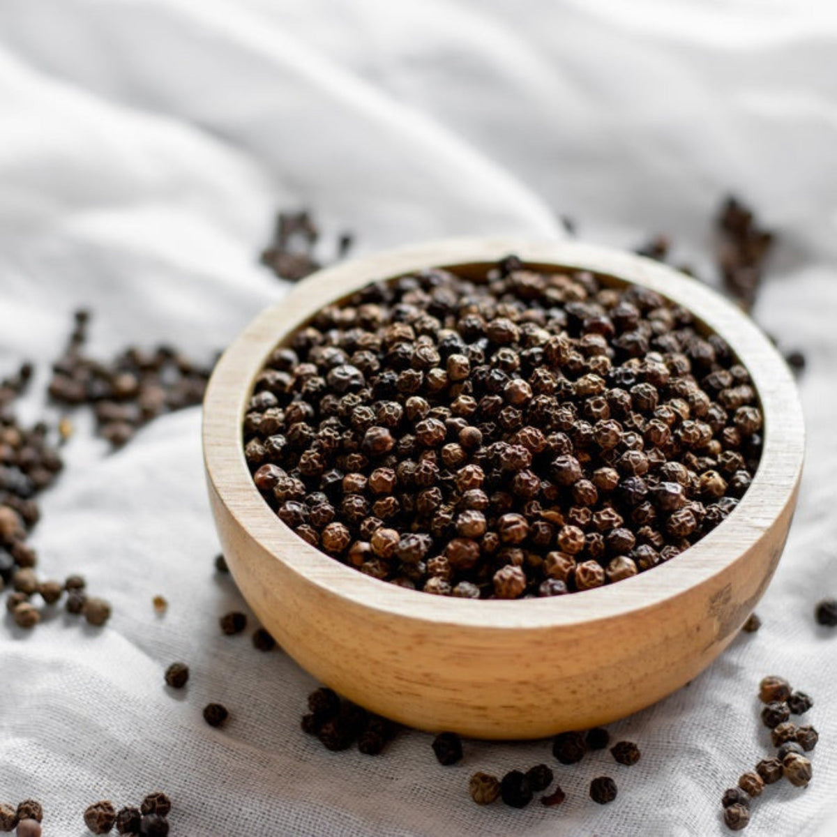 A small wooden bowl filled to the brim with whole black peppercorns, resting on a textured white cloth with loose peppercorns scattered around the base.