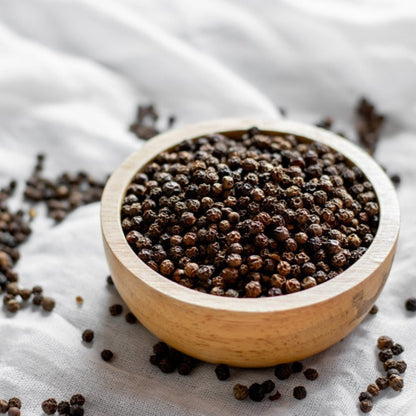 A small wooden bowl filled to the brim with whole black peppercorns, resting on a textured white cloth with loose peppercorns scattered around the base.