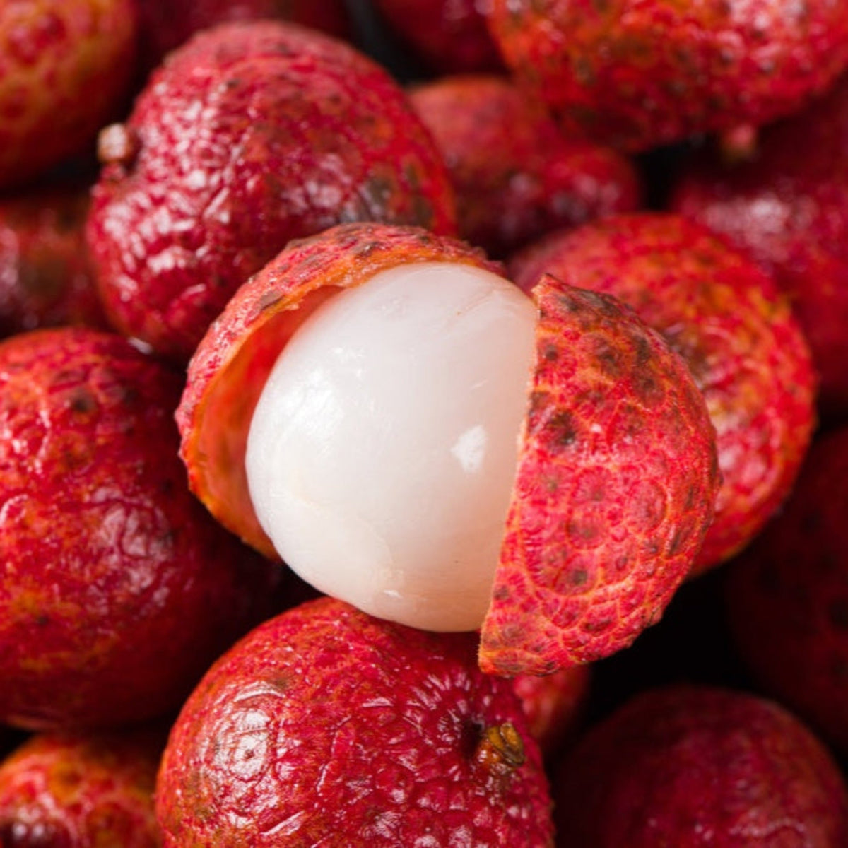 Partially peeled red lychee showing translucent white flesh among whole lychee fruits.