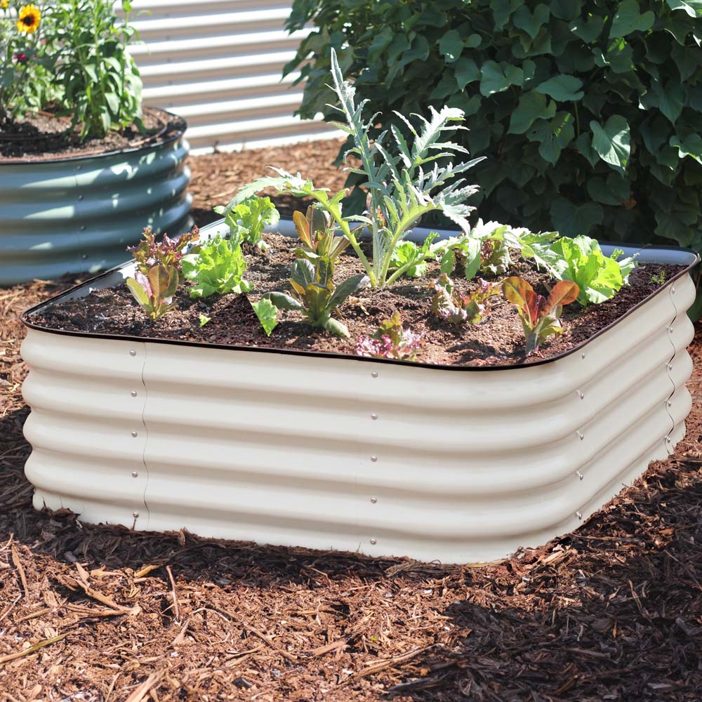 The 15-inch tall small corrugated metal raised garden bed in a light clay color, filled with various colorful flowers including pink, yellow, and purple, set in a garden bed with mulch and other plants.