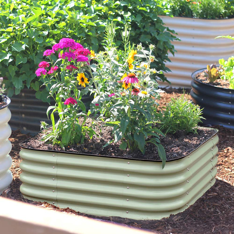 The 15-inch tall small corrugated metal raised garden bed in a mist green color, filled with various colorful flowers including pink, yellow, and purple, set in a garden bed with mulch and other plants.