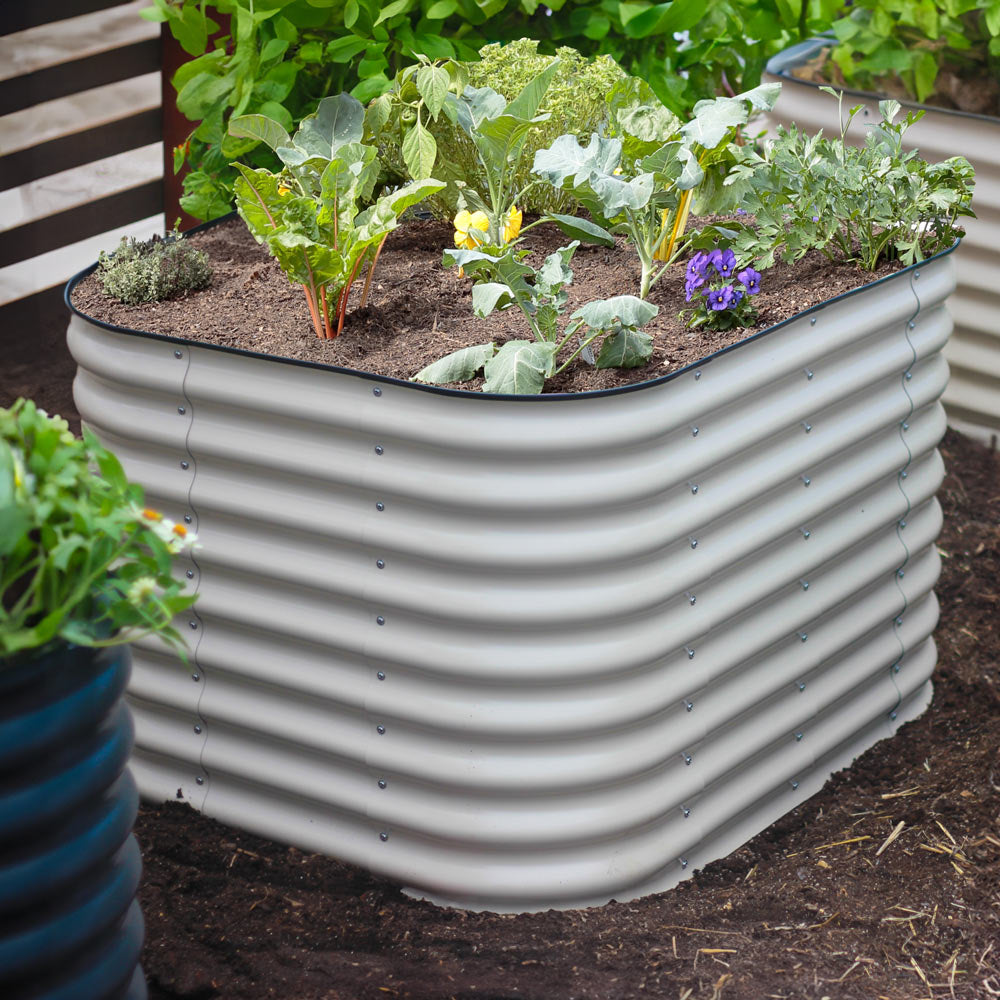 Light clay 29-inch tall small corrugated metal raised garden bed filled with various green plants and a small purple flower, set in a garden bed with soil.