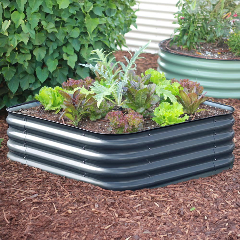 The 15-inch tall small corrugated metal raised garden bed in a slate gray color, filled with various colorful flowers including pink, yellow, and purple, set in a garden bed with mulch and other plants.