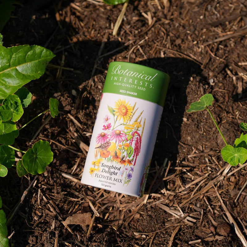 Botanical Interests Songbird Delight Flower Mix Seed Shaker canister on soil with plants around