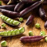 Purple Sugar Magnolia snap pea pods scattered on a wooden board, some split open to reveal rows of plump bright green peas inside.