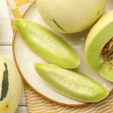 Whole and sliced honeydew melons with pale yellow-green skin and light green flesh, some seeded wedges peeled and others showing seed cavities, arranged on a plate atop a yellow-checked cloth.