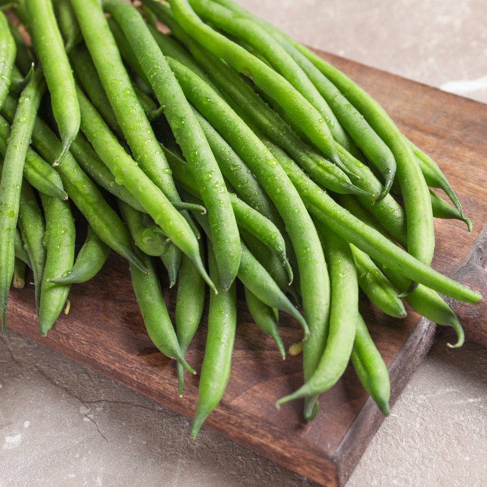 Freshly harvested Tavera filet bush beans, slender and glossy with visible water droplets, piled on a dark wooden board against a stone surface.