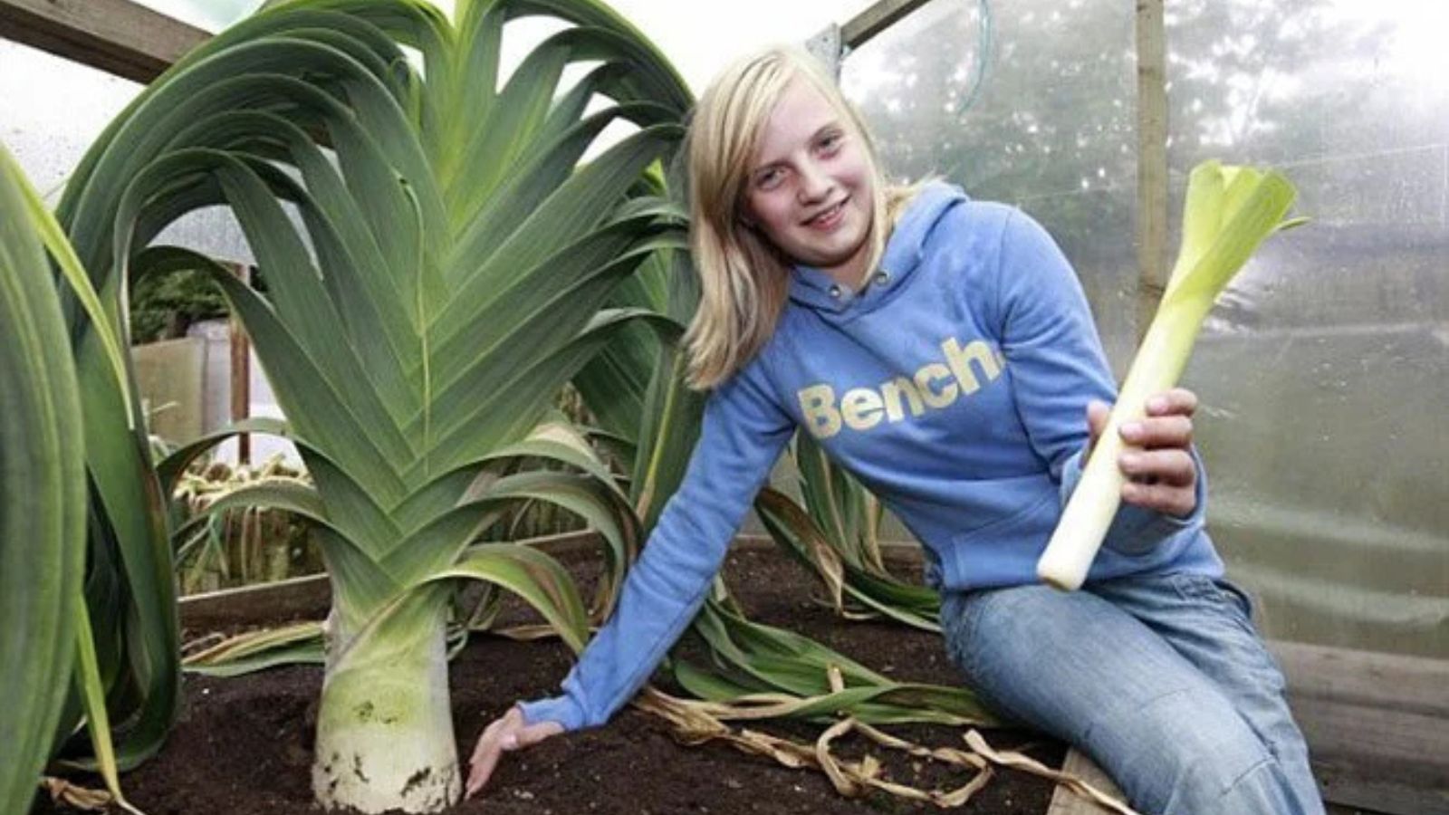 A girl sitting next to a giant leek while holding a normal-sized leek while inside a covered garden