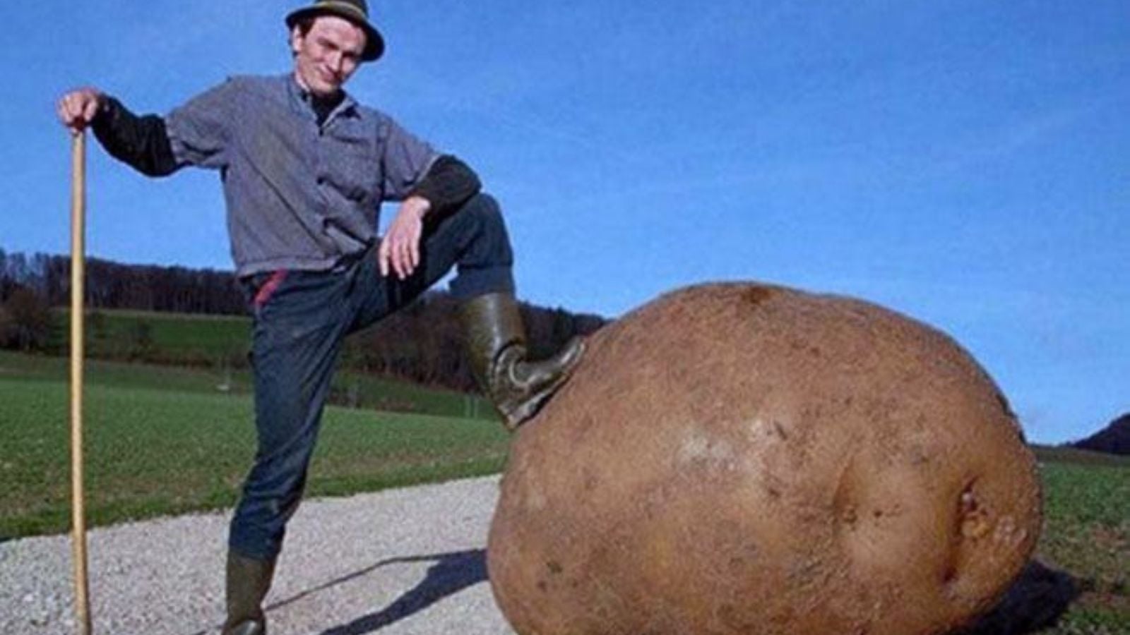A person standing beside a giant potato having hist foot on it as if it's a boulder with a field in the background