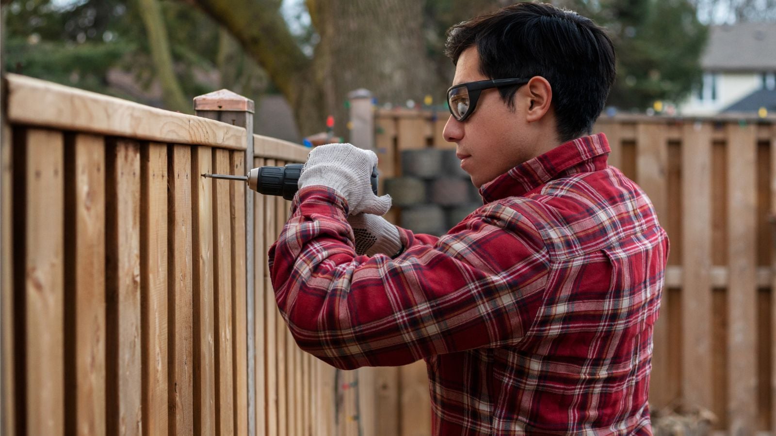 A man using a drill to work on a fence with a gate made of wood somewhere shady