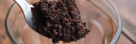 A metal spoon holds a clump of damp, dark brown organic matter with a granular texture, resting above a transparent glass bowl on a wooden surface.