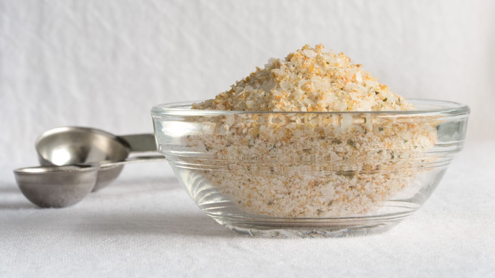 A glass bowl with salt made with Allium sativum with steel spoons in the background on a white textured surface