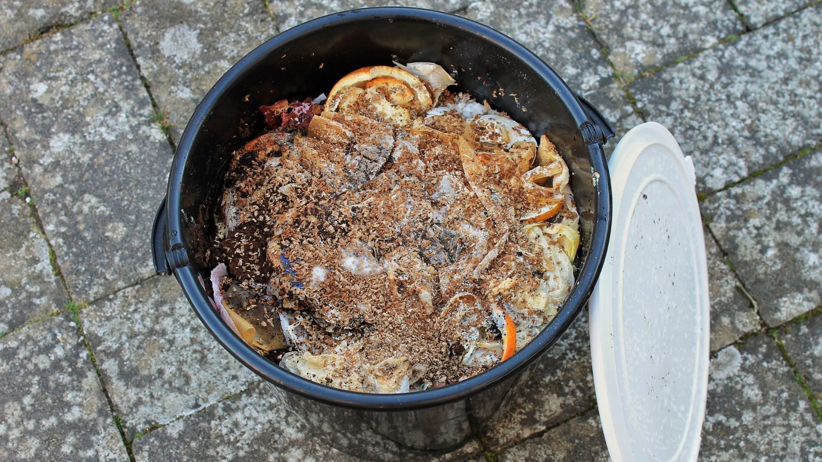 A close-up and overhead shot of a black colored bucket, filled with bran and food scraps, showcasing how to make a DIY bokashi