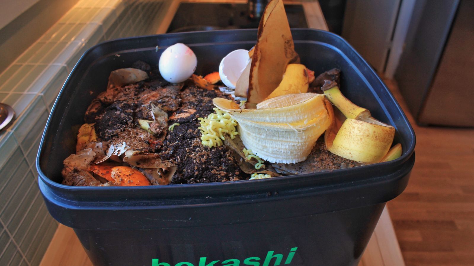 A close-up shot of a black bin filled with food scraps, placed on a kitchen table indoors