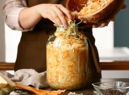 A person wearing a brown apron using bare hands to place shredded vegetables in a large glass jar placed on a wooden table