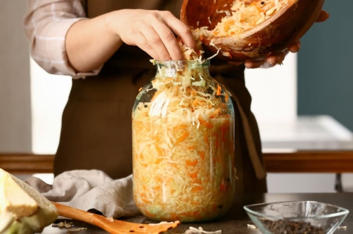 A person wearing a brown apron using bare hands to place shredded vegetables in a large glass jar placed on a wooden table
