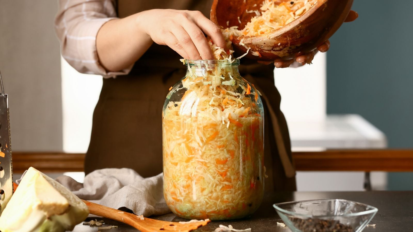 A person wearing a brown apron using bare hands to place shredded vegetables in a large glass jar placed on a wooden table
