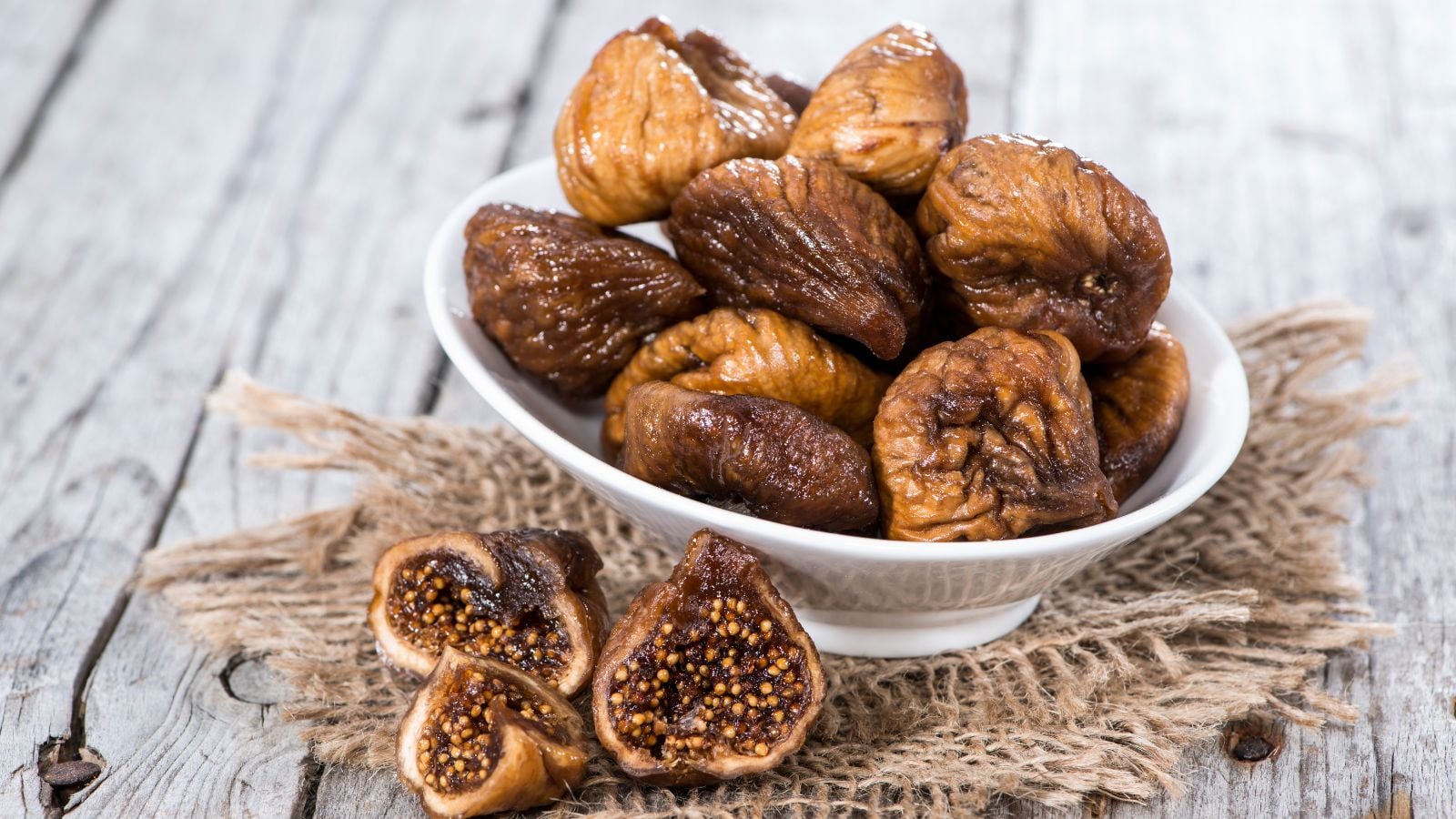 A close-up shot of a bowl filled with a small pile of dried fruits, placed on a piece of sack, on a wooden surface, in a well lit area