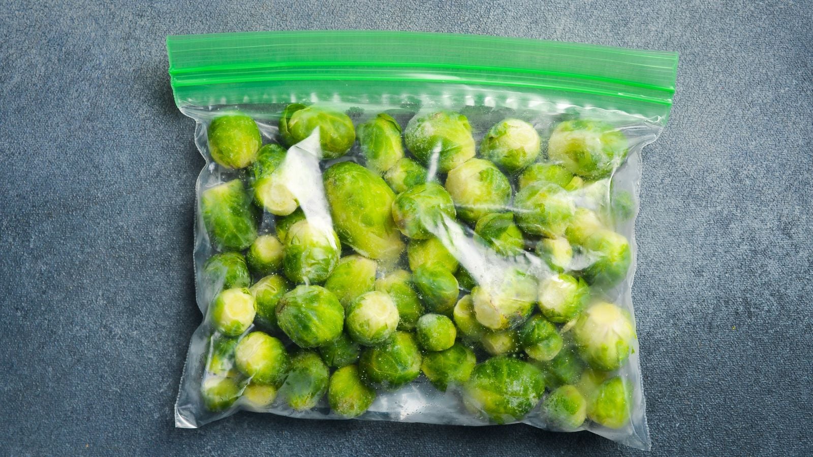 A close-up and overhead shot of a plastic bag filled with frozen heads of a crop, placed on top of a gray marble surface, all situated in a well lit area indoors