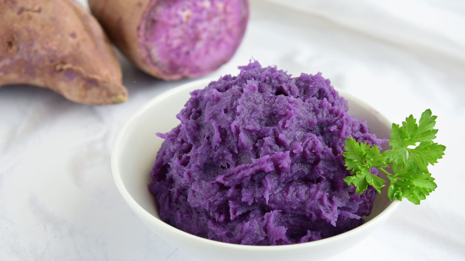 A shot of a small plate filled with a mash of purple yam, alongside raw, sliced yams, all placed on a white surface covered with tablecloth