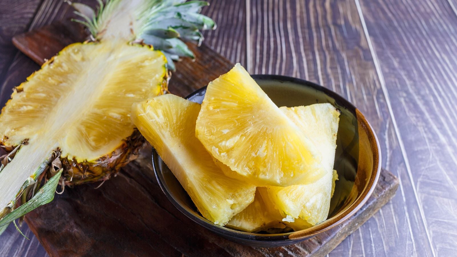 A sliched Ananas comosus fruit, showing bright yellow flesh while on a wooden plate placed on a dark wooden table