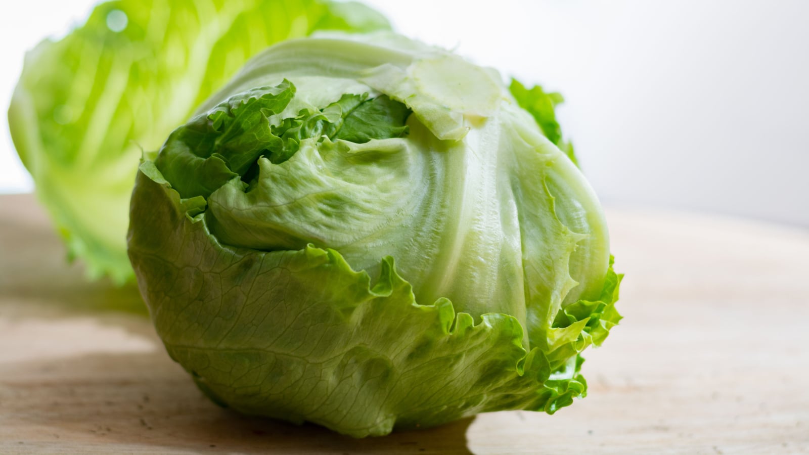 Close-up of a dense Lactuca sativa 'Iceberg' head with broad, curly-edged green leaves, sitting on a wooden countertop indoors.