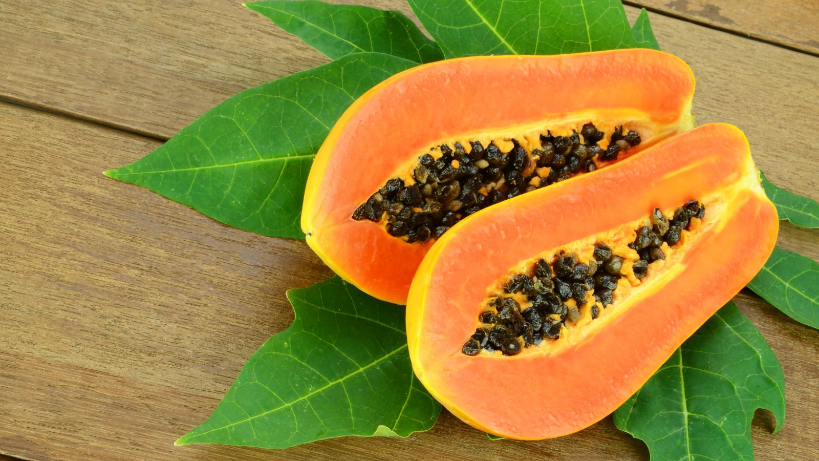 A shot of a ripe fruit with orange-colored flesh and black seeds, placed on top of its leave and a wooden surface indoors.