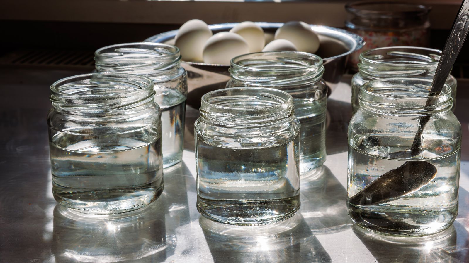 Jars filled with water with a bowl of eggs in the background, appearing to be near a window letting in bright light