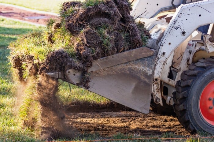 A view of a small bulldozer removing grass from a yard. The bulldozer appears worn and is white with thick rubber wheels and red rims. The surrounding green grass is still attached and rooted in dark brown soil. Clumps of earth can be seen being picked up by the bulldozer for pool installation preparation.