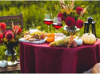 A beautiful outdoor table is set for Thanksgiving, with a burgundy table cloth, wine-red dahlias in an arrangement, and pumpkins and garden greenery.