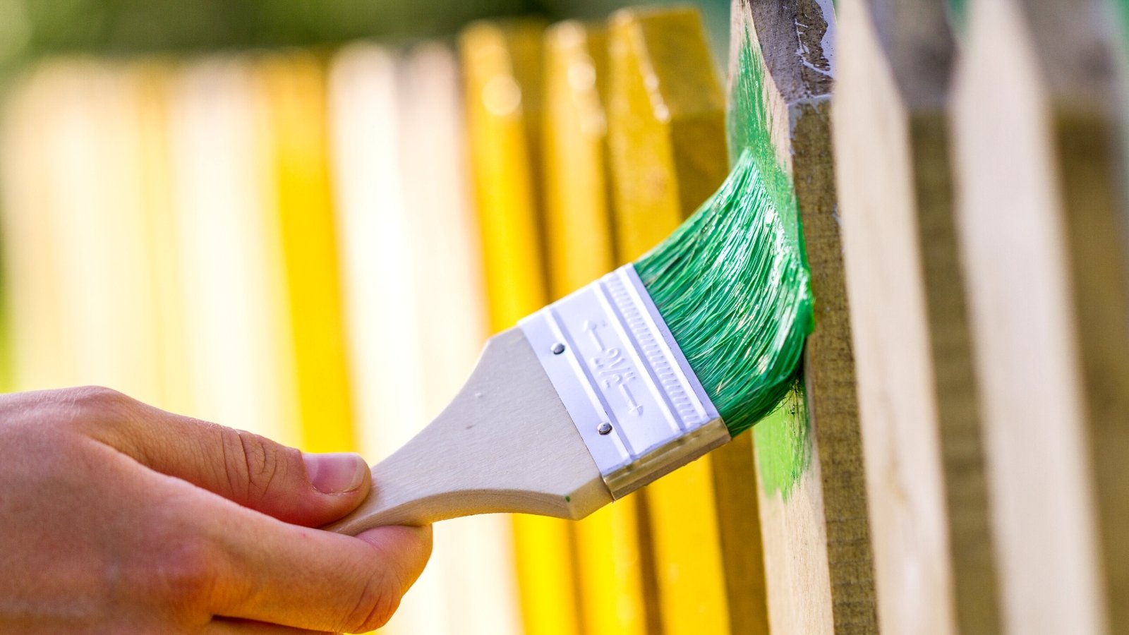 Painting fence. Close-up of a gardener's hand painting a wooden fence with a brush. Part of the fence is bright yellow, and the brush paints in bright green.