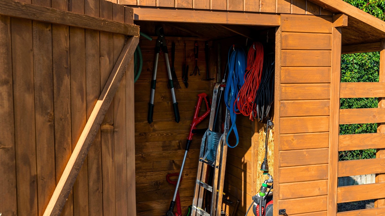 Close-up of a wooden garden shed with various gardening things such as secateurs, garden shears, lawn mower, stepladder, spades, rakes, hoses and others.