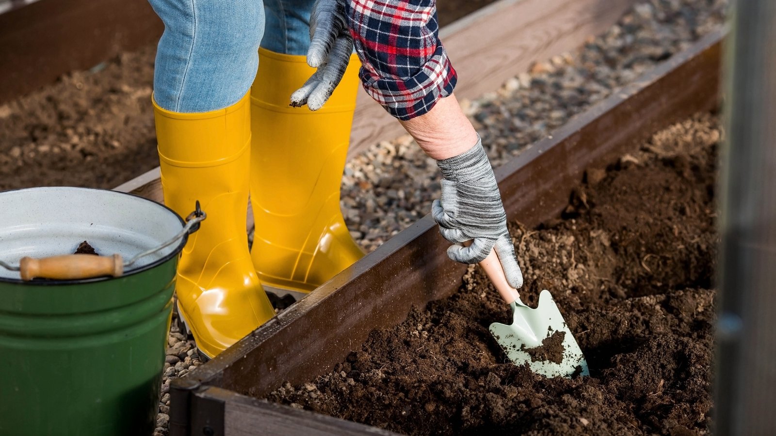 Close-up of a female gardener in jeans, high yellow rubber boots preparing the soil in a raised bed using a trowel.