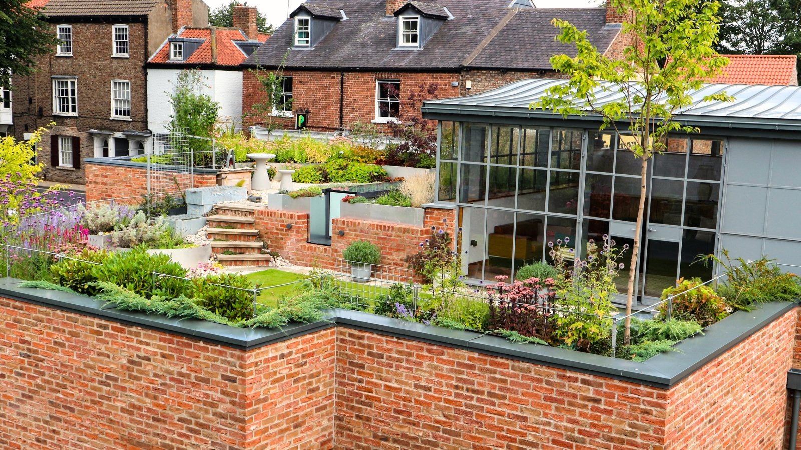 View of a two-story brick house with a stunning flowering rooftop garden including Orpine, Echinops, Common juniper, Verbena, and other perennials.