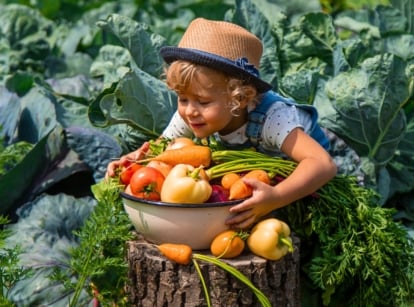 A girl with curly white hair wearing a straw hat embraces a large bowl brimming with freshly picked vegetables, including bell peppers, tomatoes, and carrots.