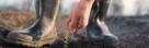 A person in sturdy boots scattering tiny grains over freshly tilled soil in an outdoor garden.