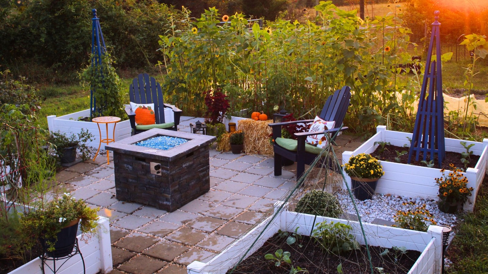 A charming patio features white wooden raised beds and planters filled with colorful flowers and vegetables, encircling a cozy seating area with chairs and a fireplace.