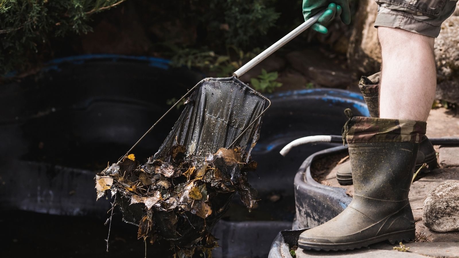 A person in rubber boots cleaning debris out of water using a black-handled net, with wet leaves and dirt visible in the netting.