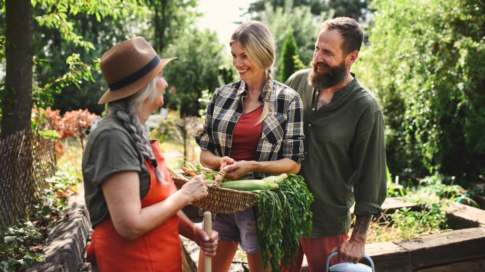 A young couple, a woman and a man, holding a basket filled with vegetables like corn, carrots, and beets, converse with an elderly woman in a gardening community.