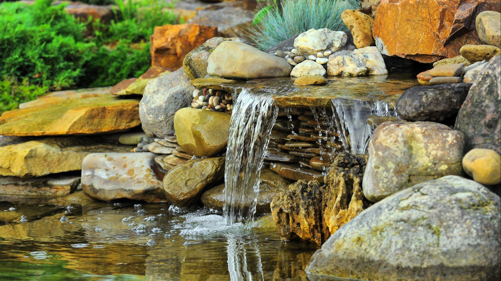 A small man-made waterfall flowing over smooth, stacked stones with plants and pebbles surrounding the water.