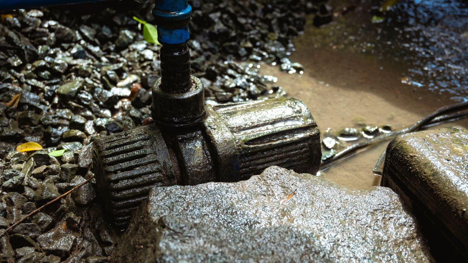A muddy water pump with a hose attached, placed on wet ground with dirt, gravel, and puddles in the background.
