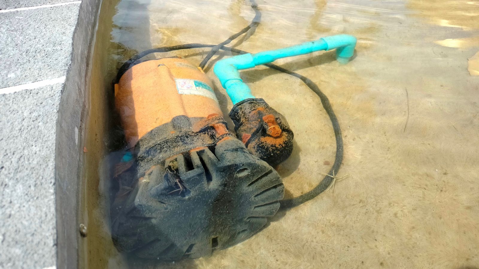 A water pump partially submerged in clear water, with a bright orange motor and blue handle visible on the surface.