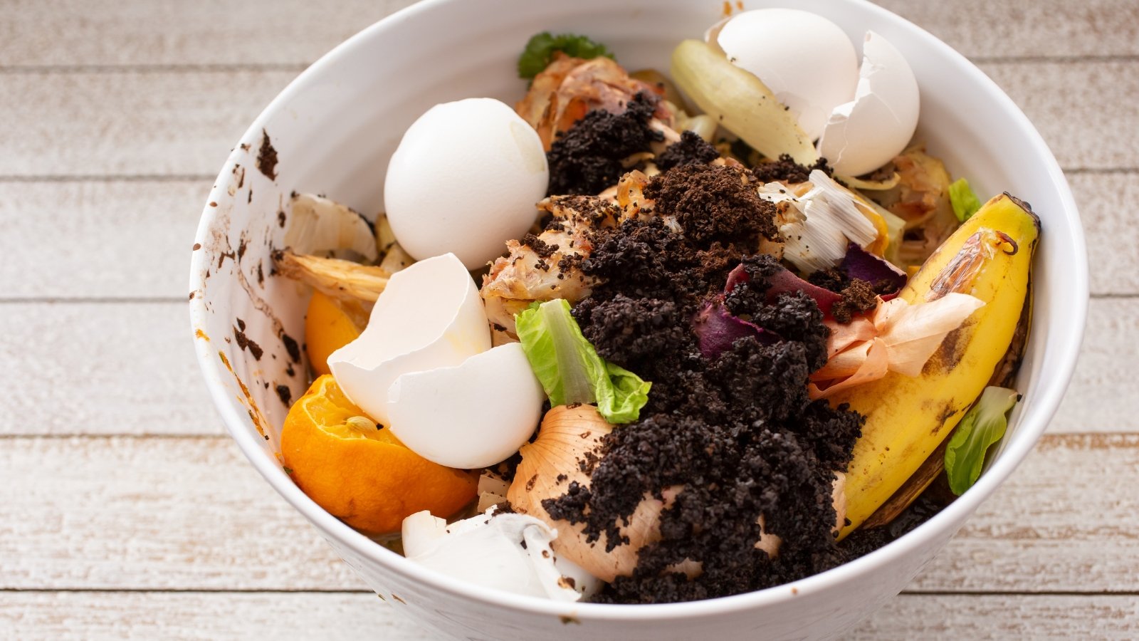 A white bowl containing egg shells, banana peels, apple cores, and other colorful food scraps on a wooden surface.